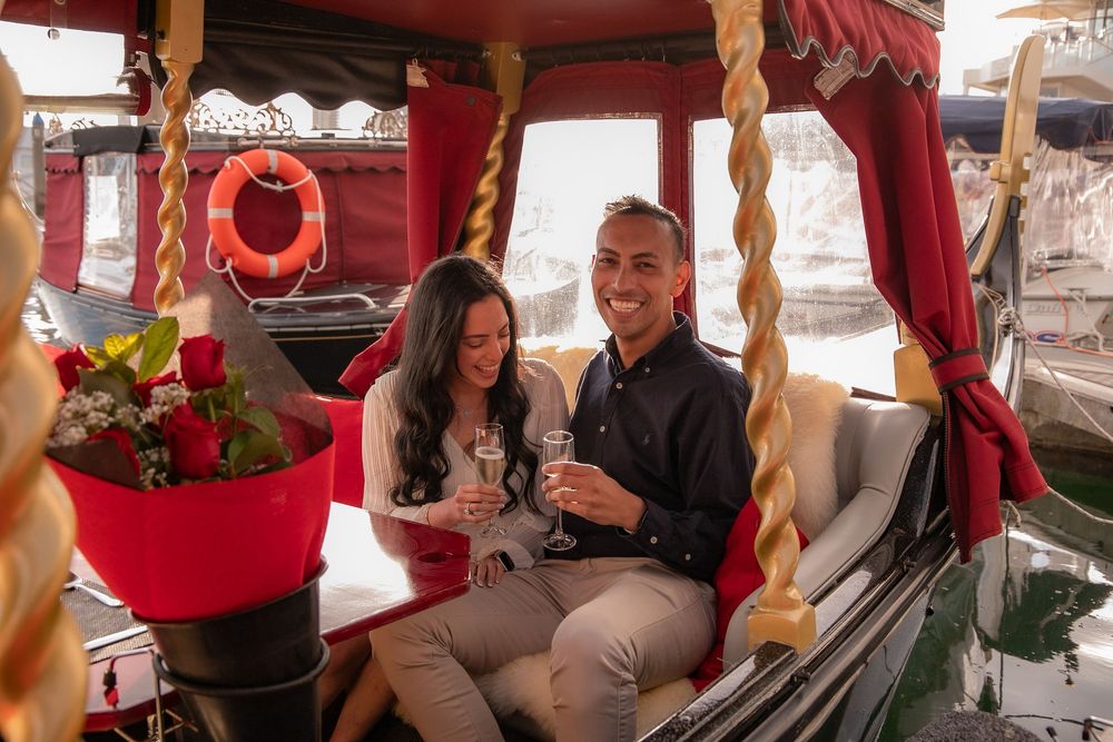 Couple smiling and holding champagne glasses on a romantic gondola boat ride with red roses.