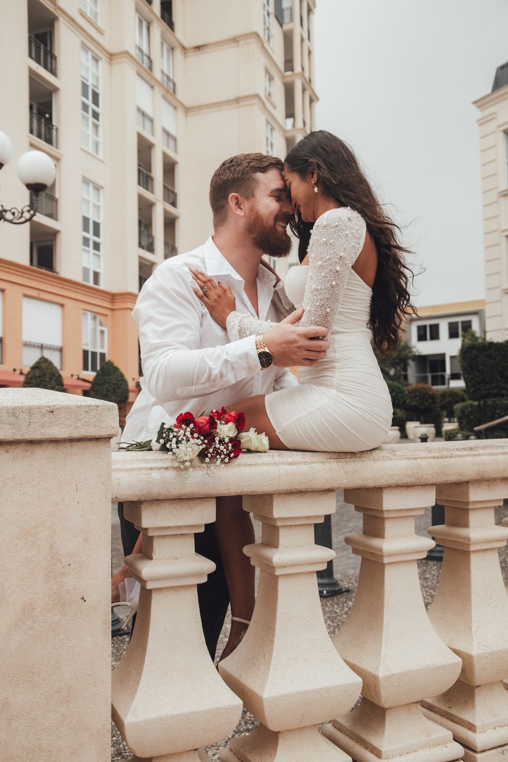 Smiling couple in white outfits sharing a romantic moment on an elegant balcony with flowers.