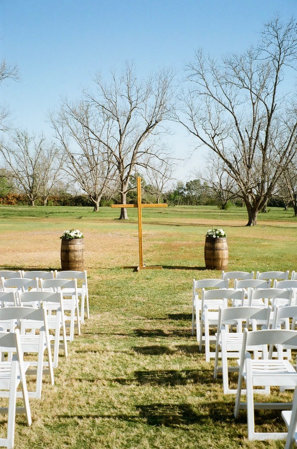 Outdoor wedding ceremony setup with white chairs, wooden cross altar, barrel flower arrangements, and bare trees under blue sky.