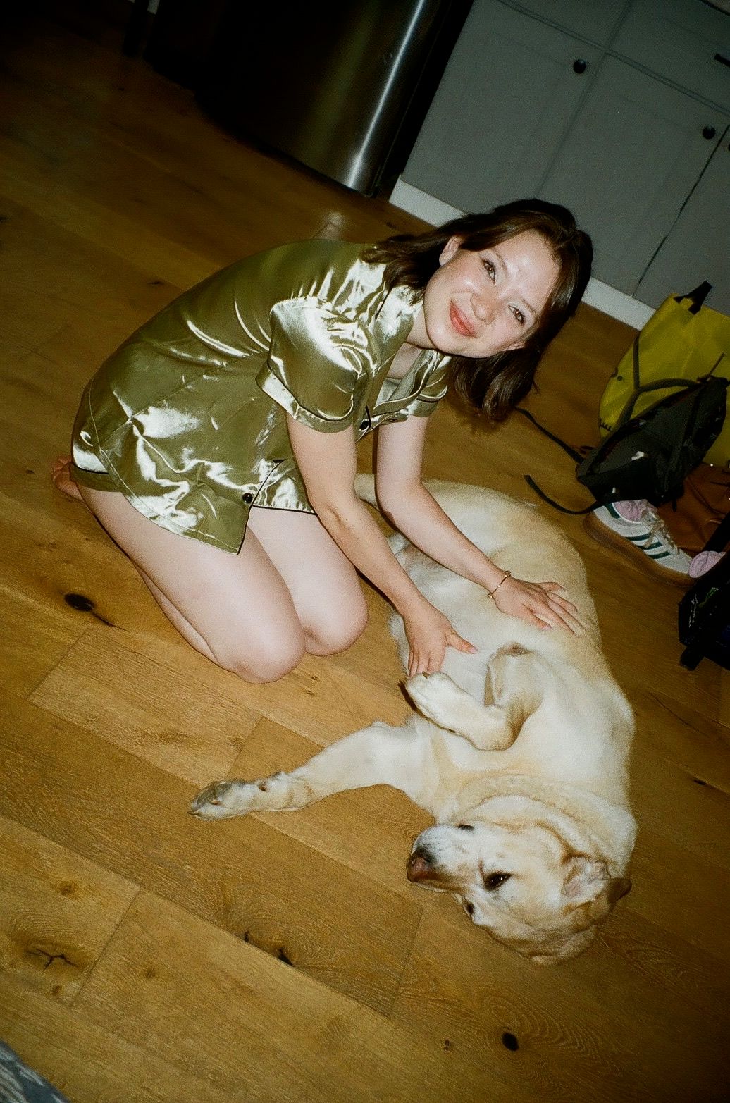 Young woman in gold satin dress kneeling on hardwood floor petting a relaxed yellow Labrador dog.