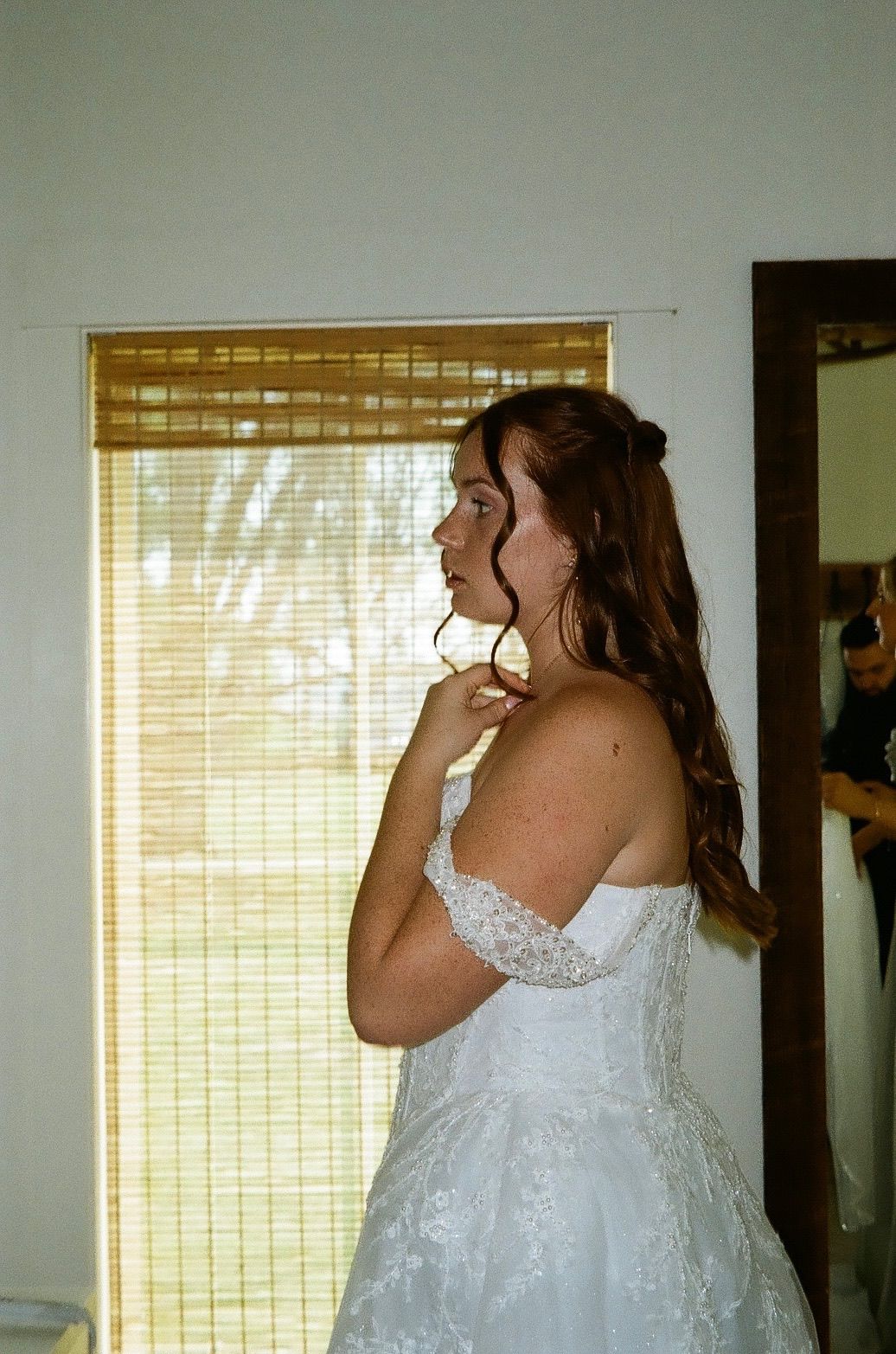 Bride in white off-shoulder lace wedding gown gazing out a window with bamboo blinds, long brown hair styled elegantly.