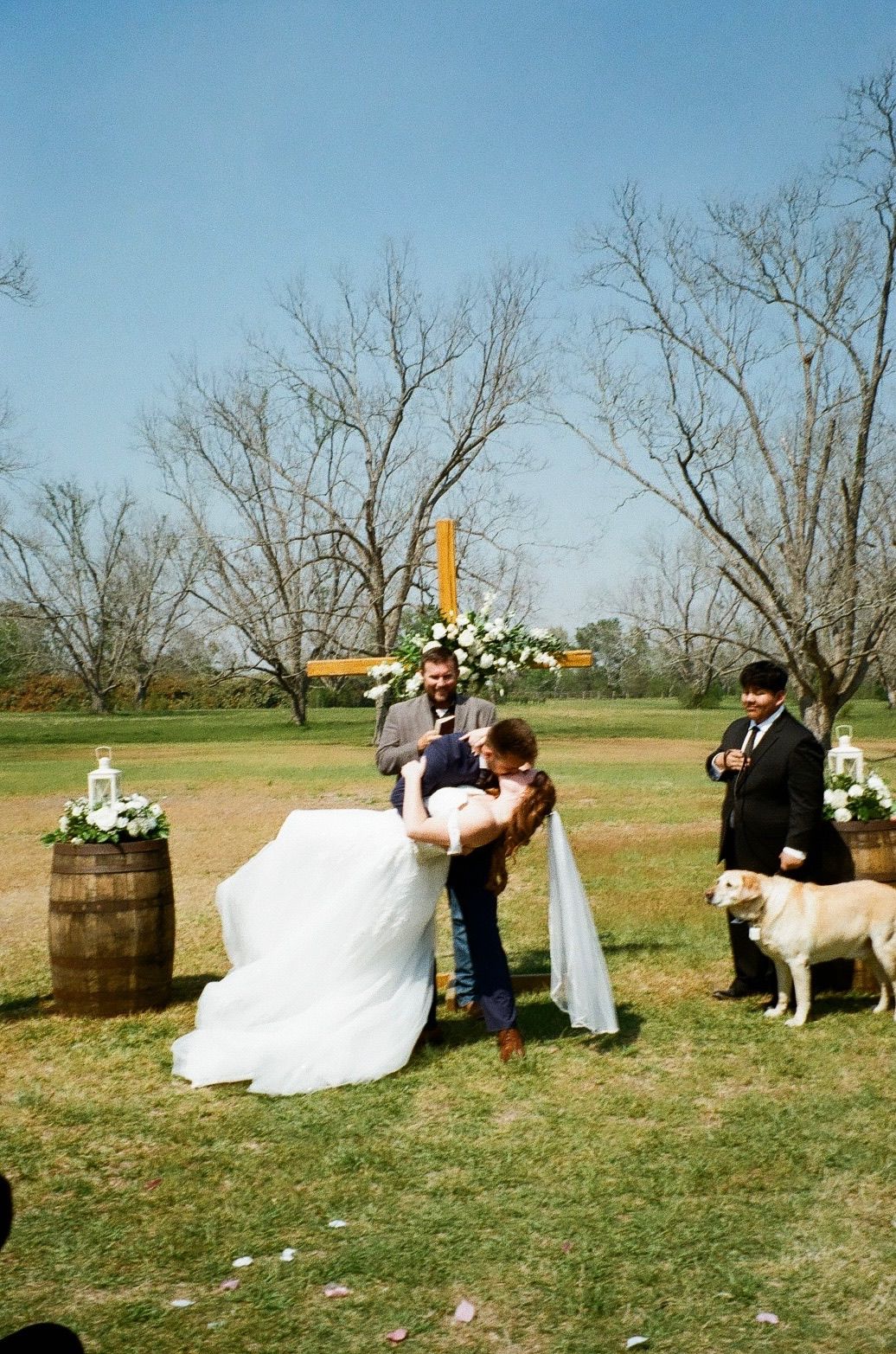 Groom lifting bride at outdoor wedding ceremony with wooden cross altar, barrel decorations, and white dog on sunny day.