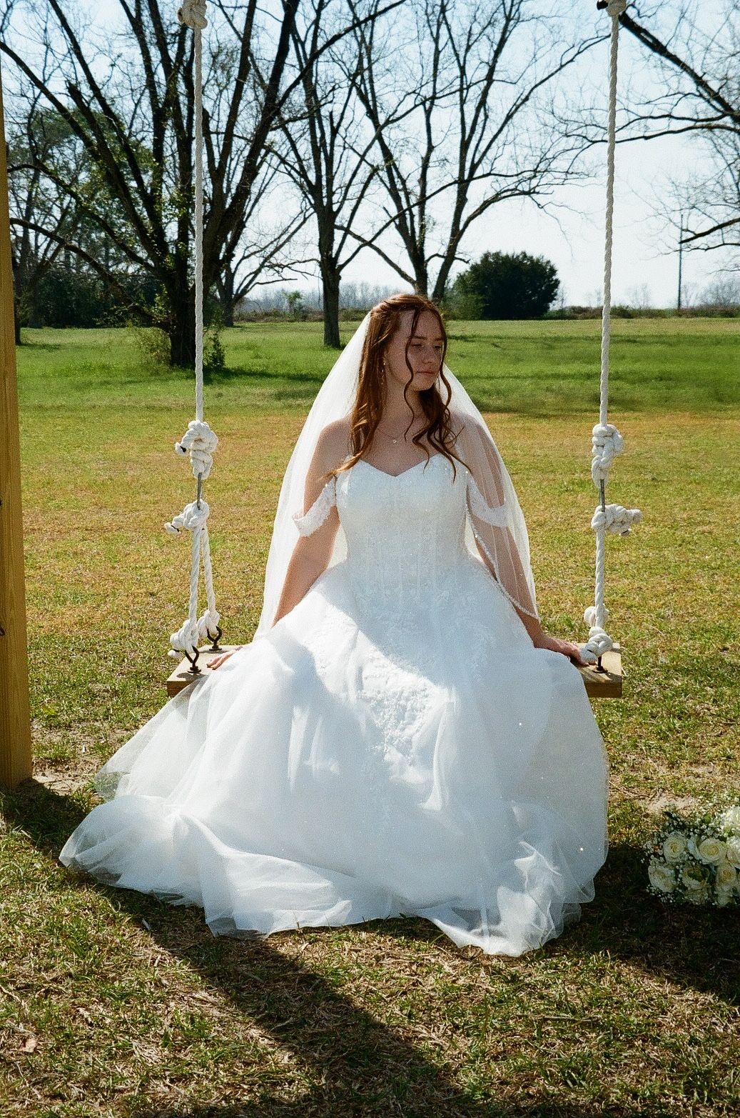 Bride in white ball gown and veil sitting on a macrame rope swing outdoors on a sunny wedding day.
