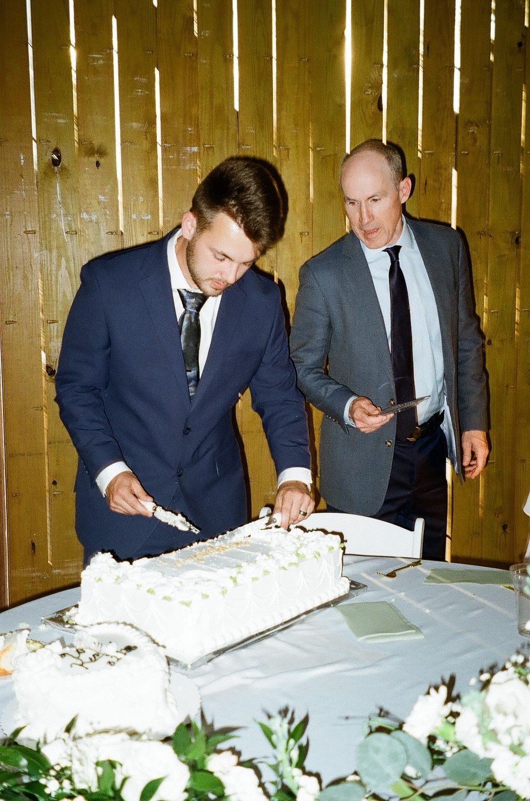 Two men in suits cutting a white wedding cake at a reception in front of a wooden wall.