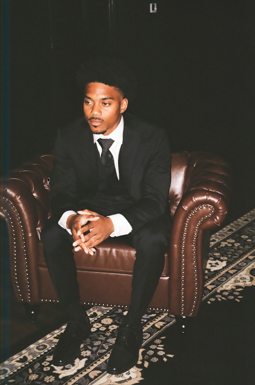Young man in black suit and tie sits confidently in brown leather armchair against dark background.