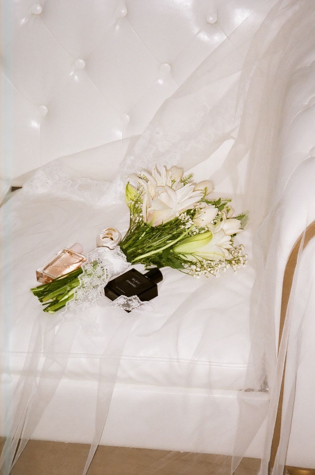 White wedding bouquet with tulips and baby's breath beside rings and perfume on tufted white chair with veil.