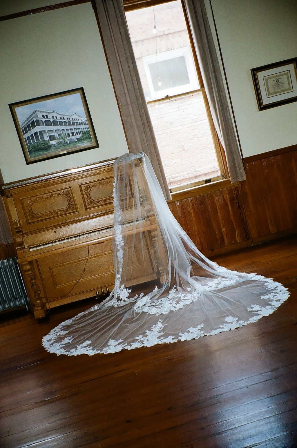 Elegant lace-trimmed cathedral wedding veil draped over a chair near a vintage upright piano on hardwood floors.
