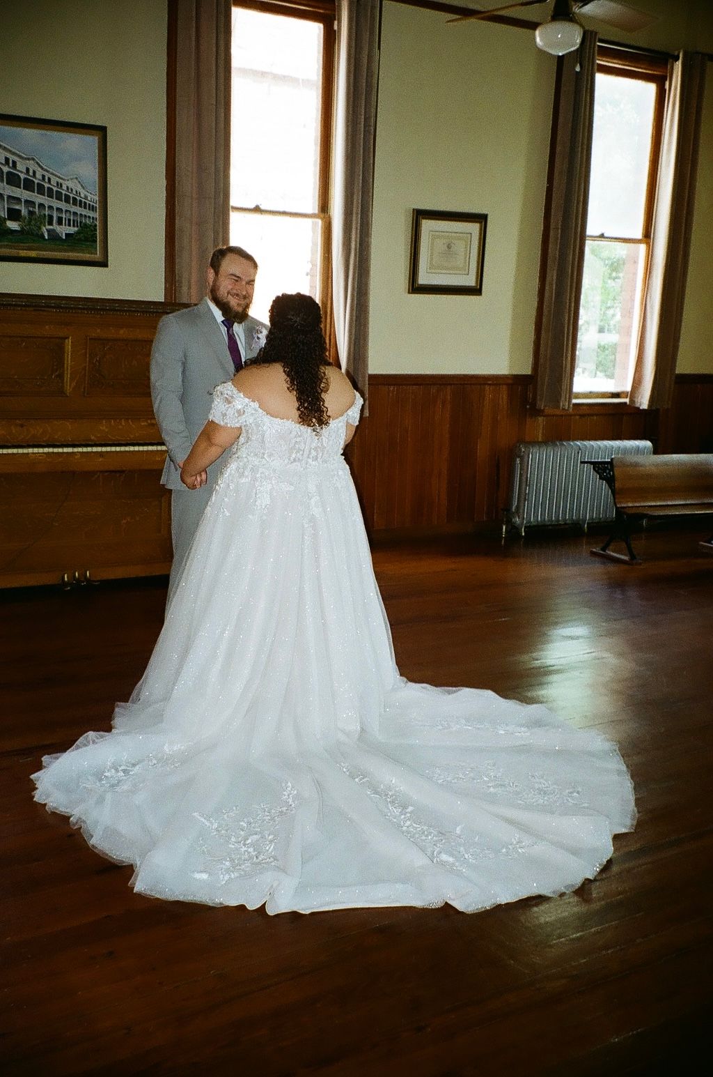 A bride in an off-shoulder white gown with a long train shares a first dance with her groom indoors.