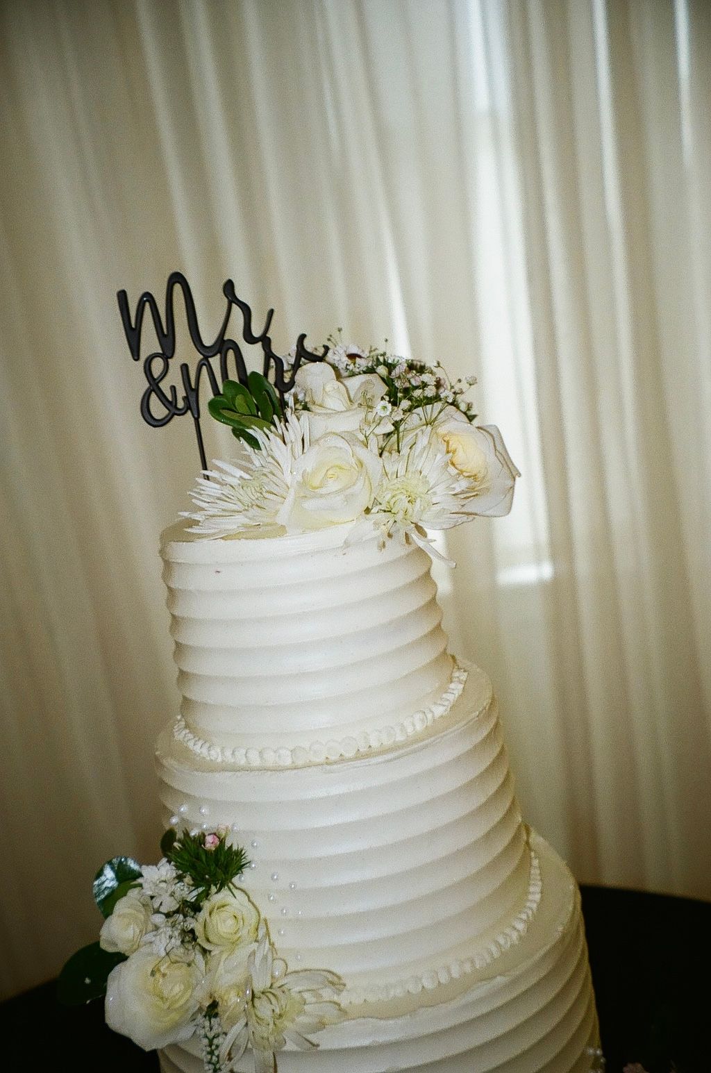 Elegant three-tier white wedding cake with Mr & Mrs topper and white floral decorations on a draped backdrop.