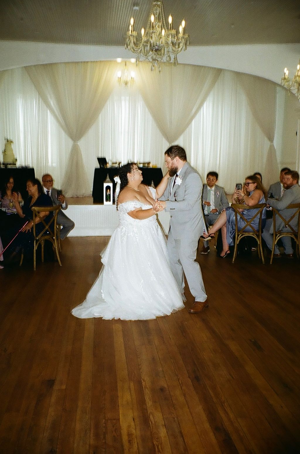 Bride and groom share their first dance on a wooden floor in an elegant wedding reception venue with chandelier lighting.
