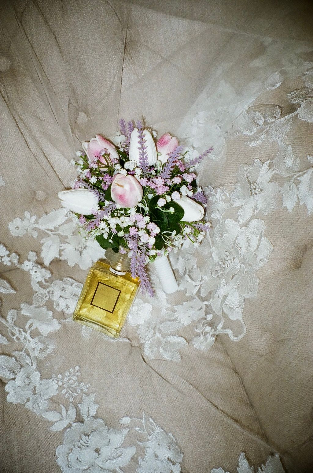 Delicate bridal bouquet with pink tulips and lavender flowers resting on lace wedding dress with perfume bottle.