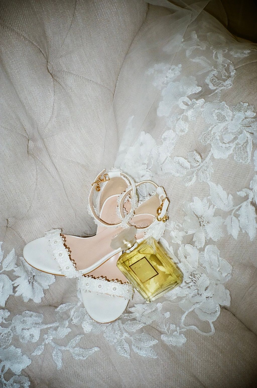 White bridal heels with gold chain detail and yellow perfume bottle on lace wedding dress.