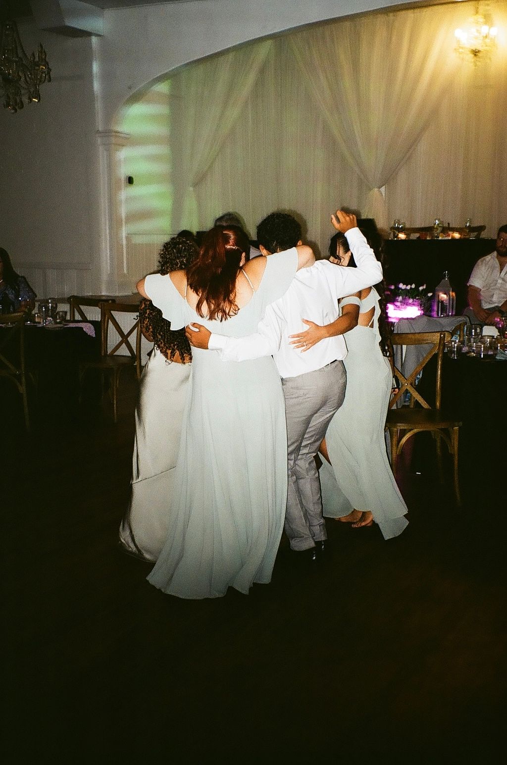 Wedding guests in white attire share a group hug on the dance floor during a festive indoor reception celebration.