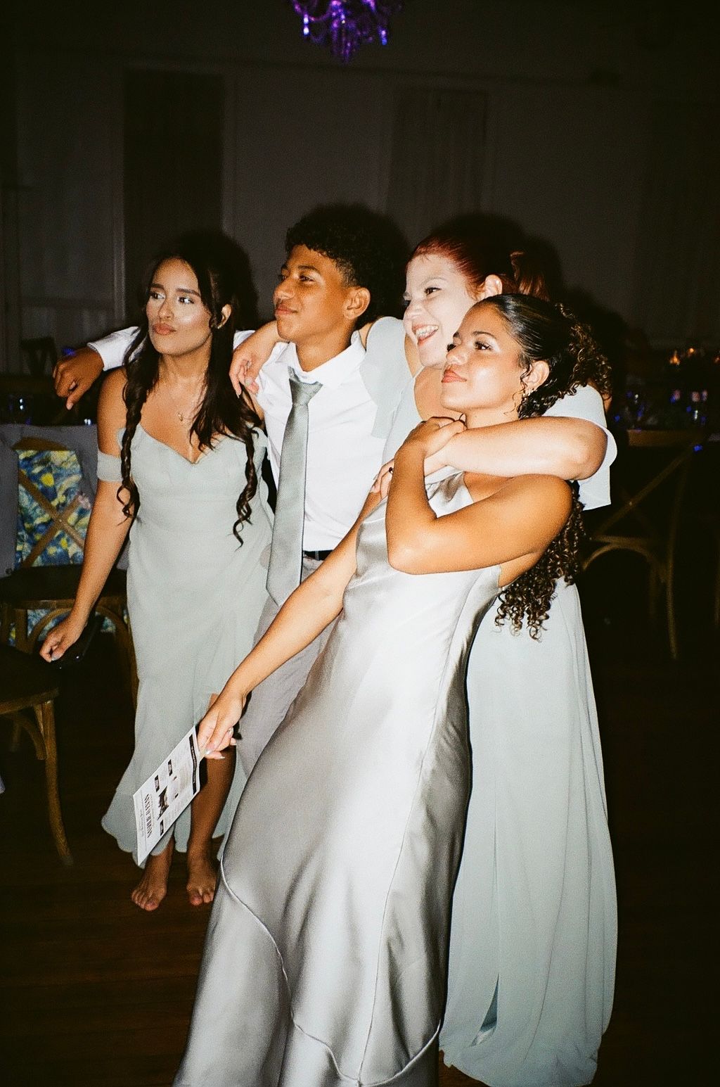 Four young wedding guests in silver dresses posing and hugging on the dance floor.