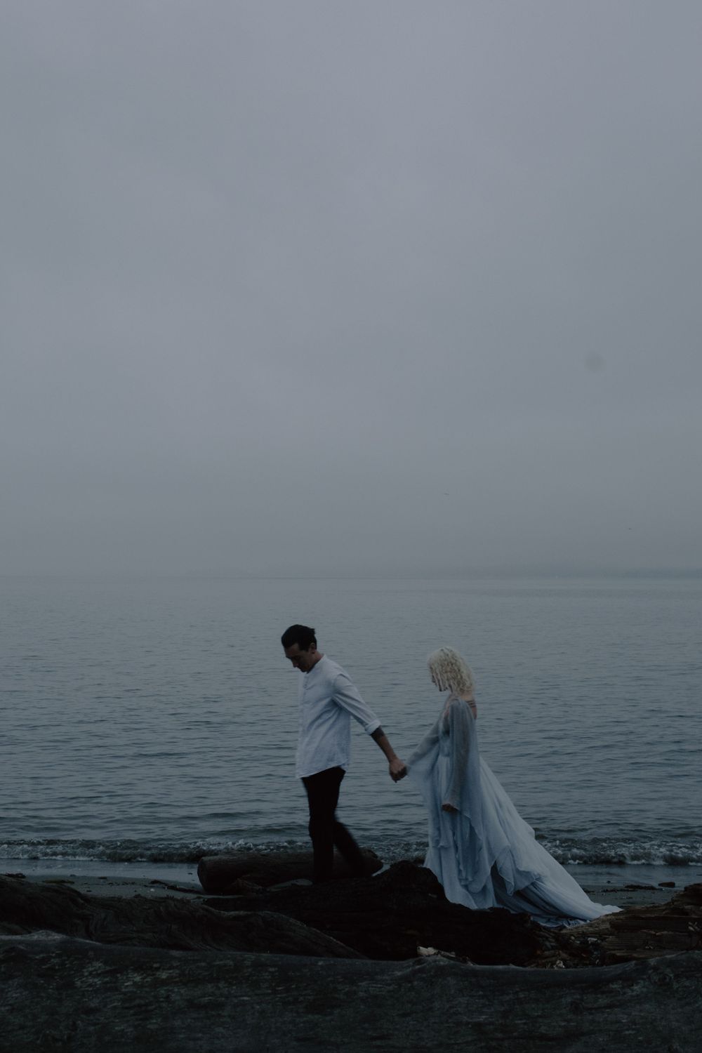 A couple walks along rocky coastal shores in wedding attire during an overcast day.