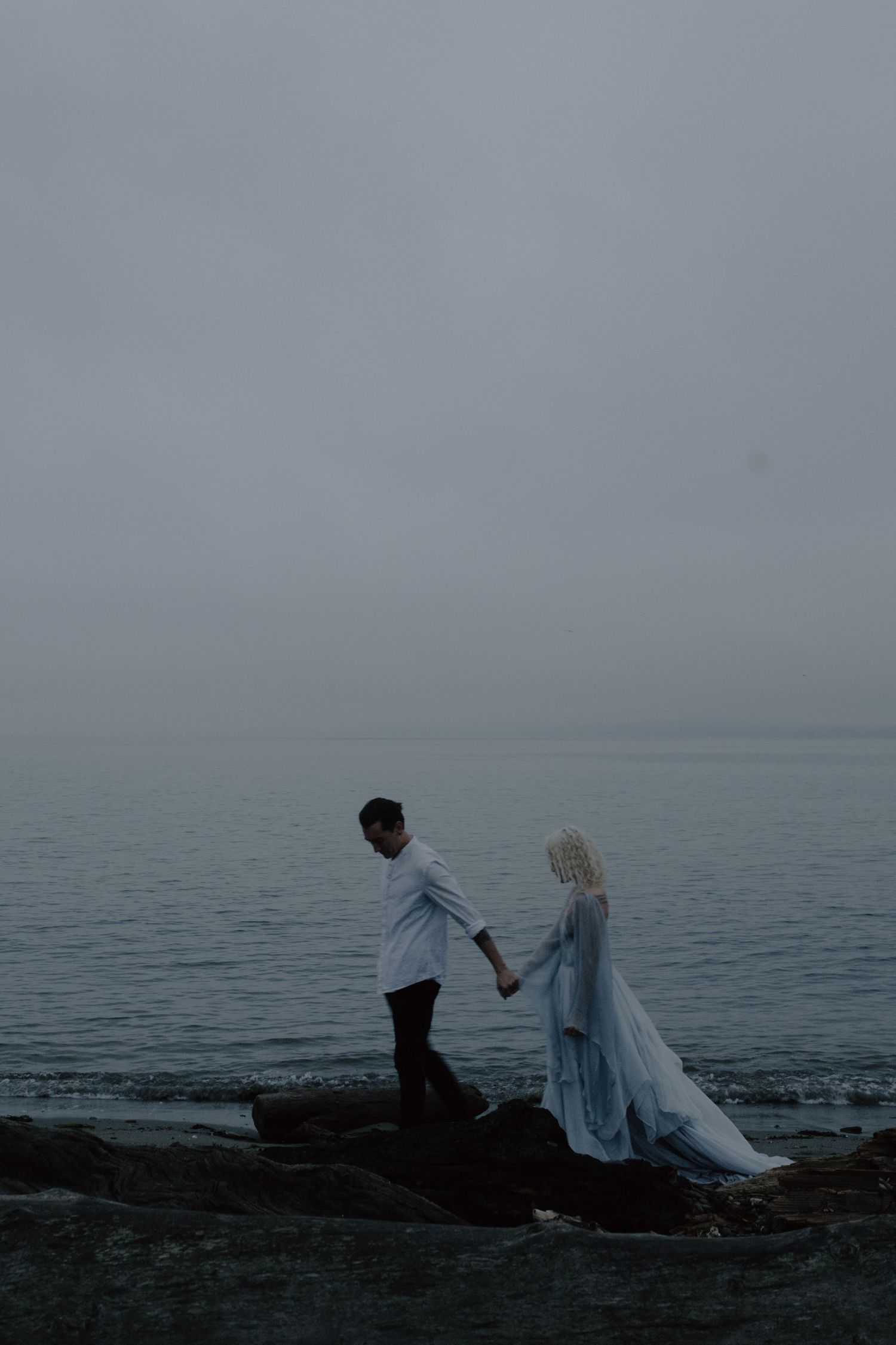 A couple walks along rocky coastal shores in wedding attire during an overcast day.