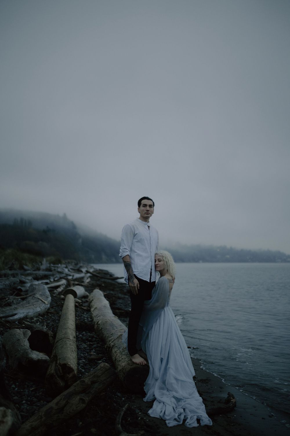 A couple in wedding attire stands on driftwood logs by the misty ocean.