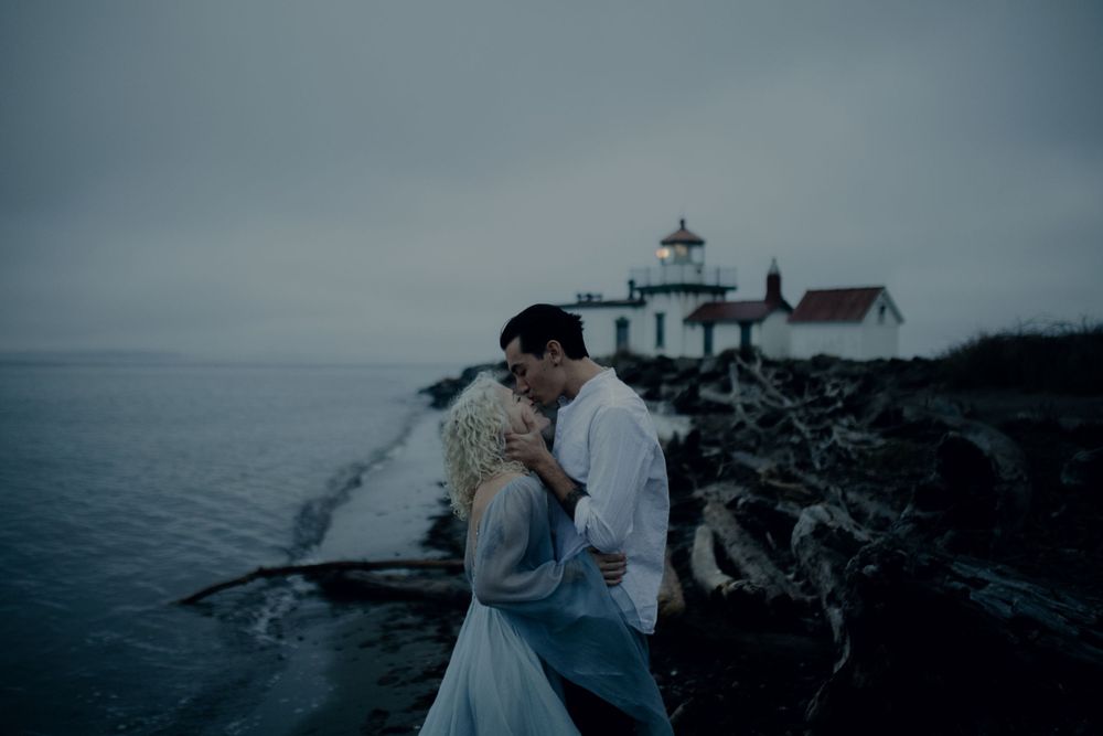 A couple embraces near a lighthouse on a foggy Pacific Northwest coastline.