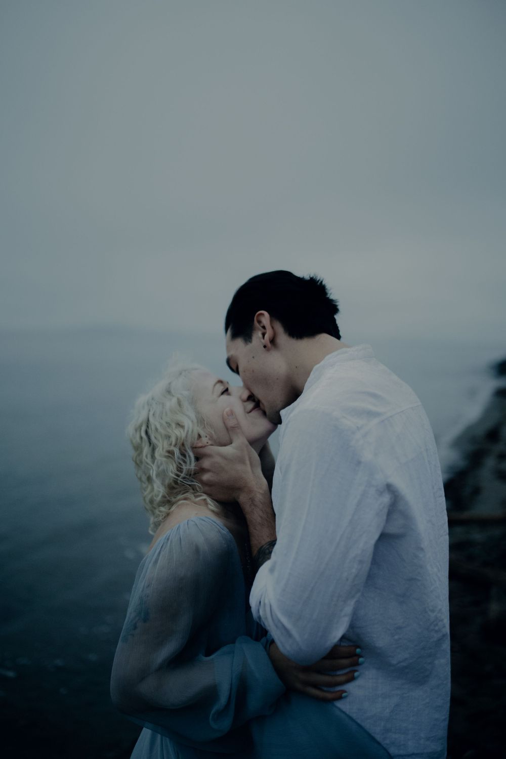 A moody seaside photoshoot with two people embracing against a misty gray backdrop.