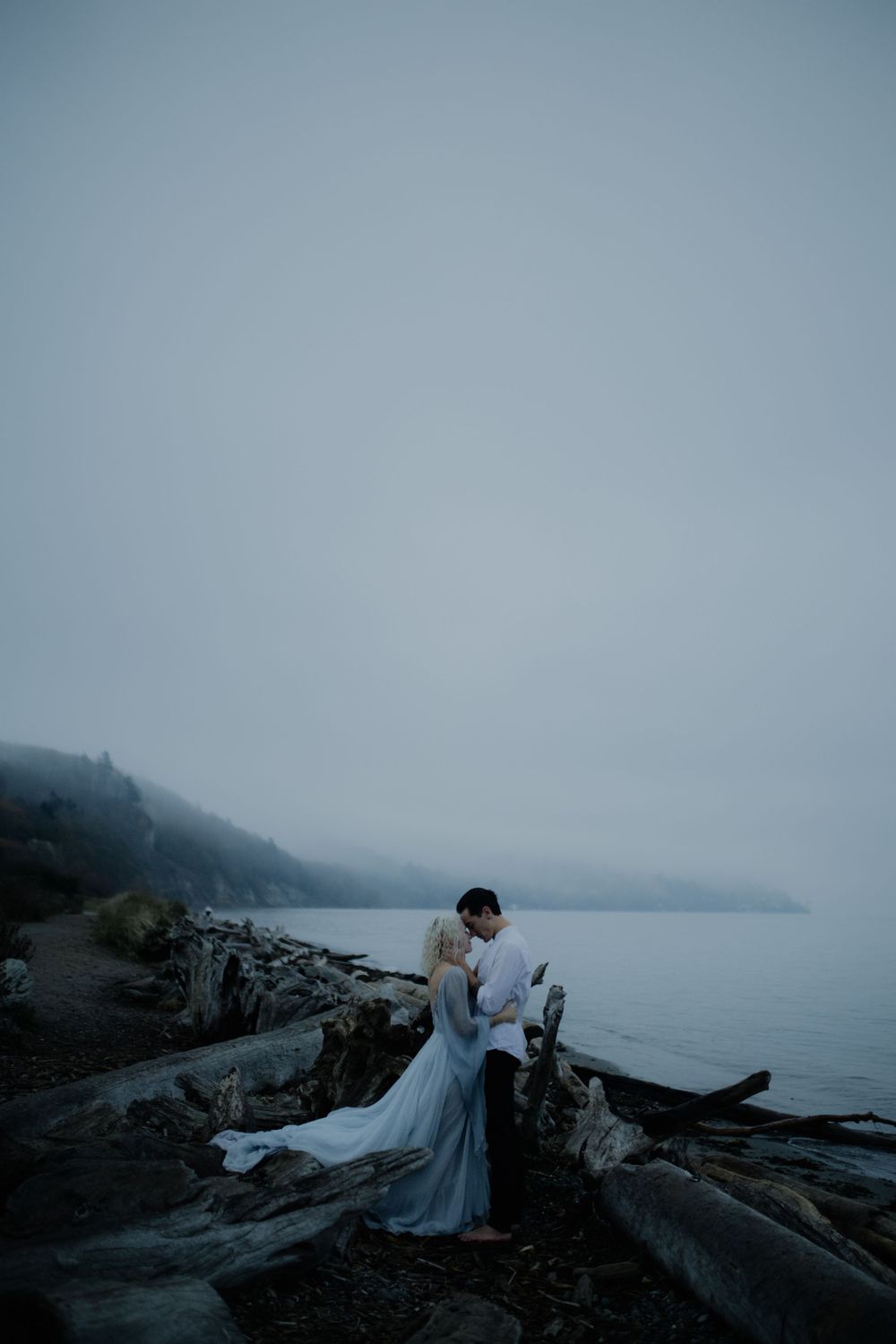 A romantic coastal photoshoot on rocky shores with a lighthouse in the foggy distance.