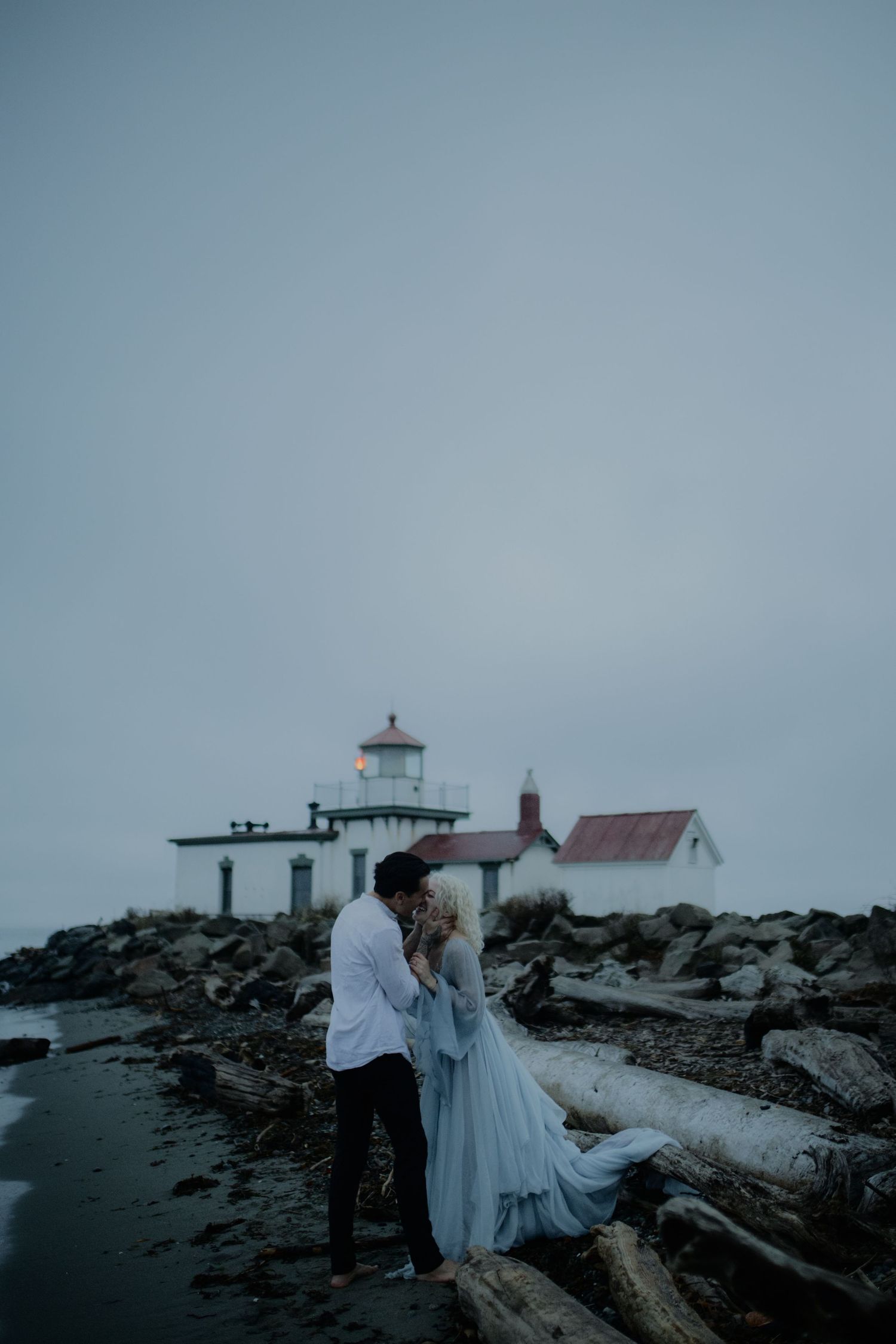 Two figures in light clothing pose near a lighthouse on rocky coastal terrain under overcast skies.