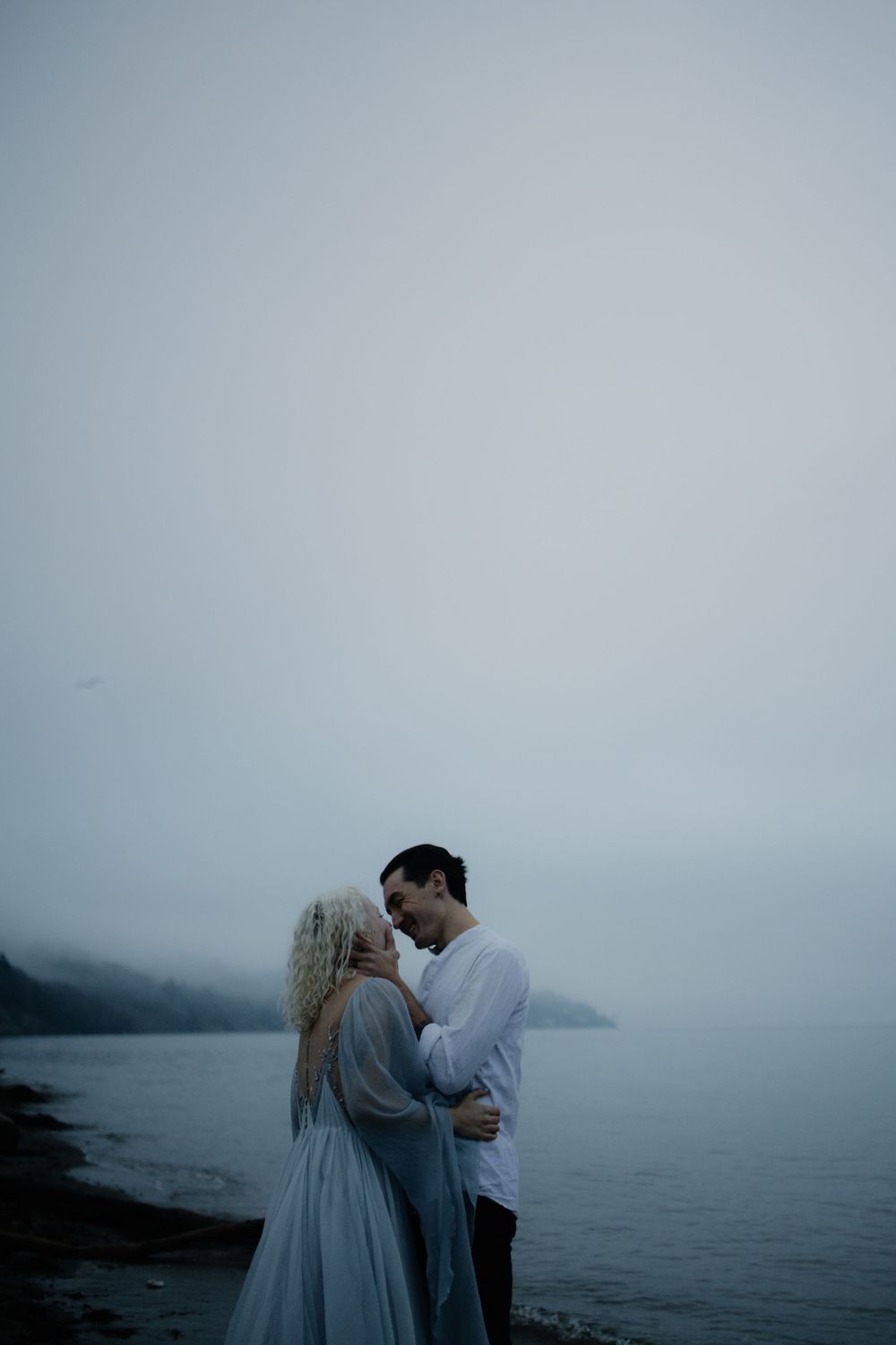 A misty oceanside portrait session with two people embracing against dramatic coastal scenery.