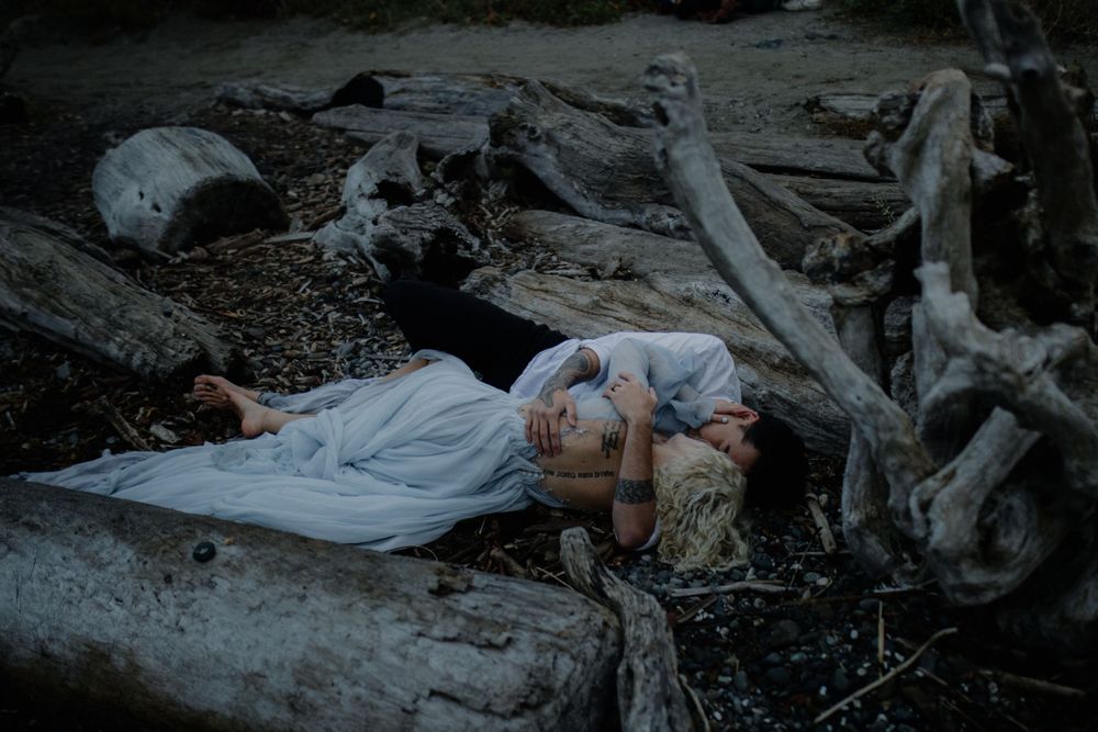 A dramatic coastal photoshoot with figures lying among driftwood on a moody beach.