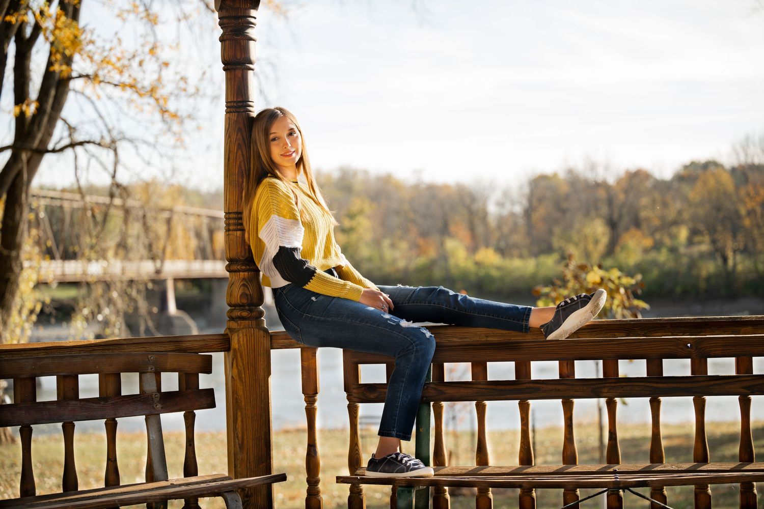 Person in casual attire sitting on a wooden deck railing during autumn with trees in background.