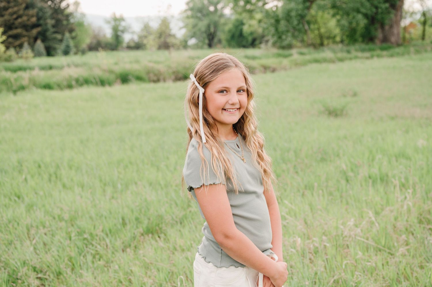 A smiling person stands in a lush green field wearing a light colored top on a sunny day.