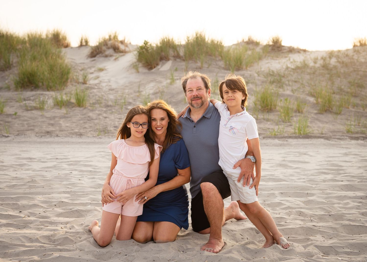 A smiling family poses together on a sandy beach with dunes in the background at sunset.