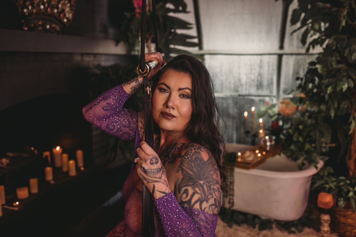 Atmospheric portrait of woman in purple lace dress with tattoos sitting among candles and greenery in moody lighting.