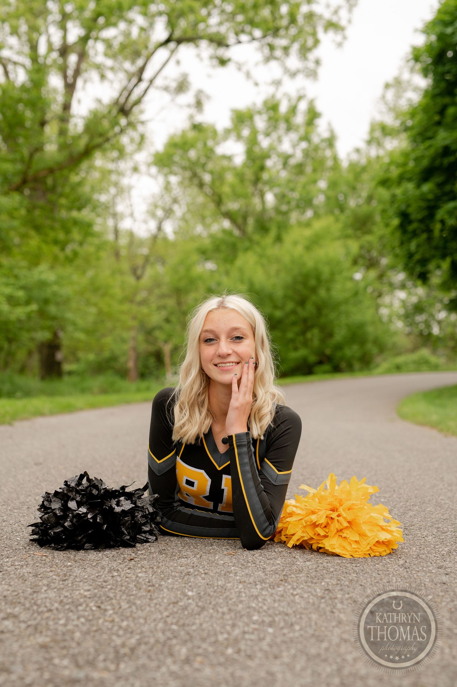 Cheerleader lies on path with black and gold pom poms against green spring background.