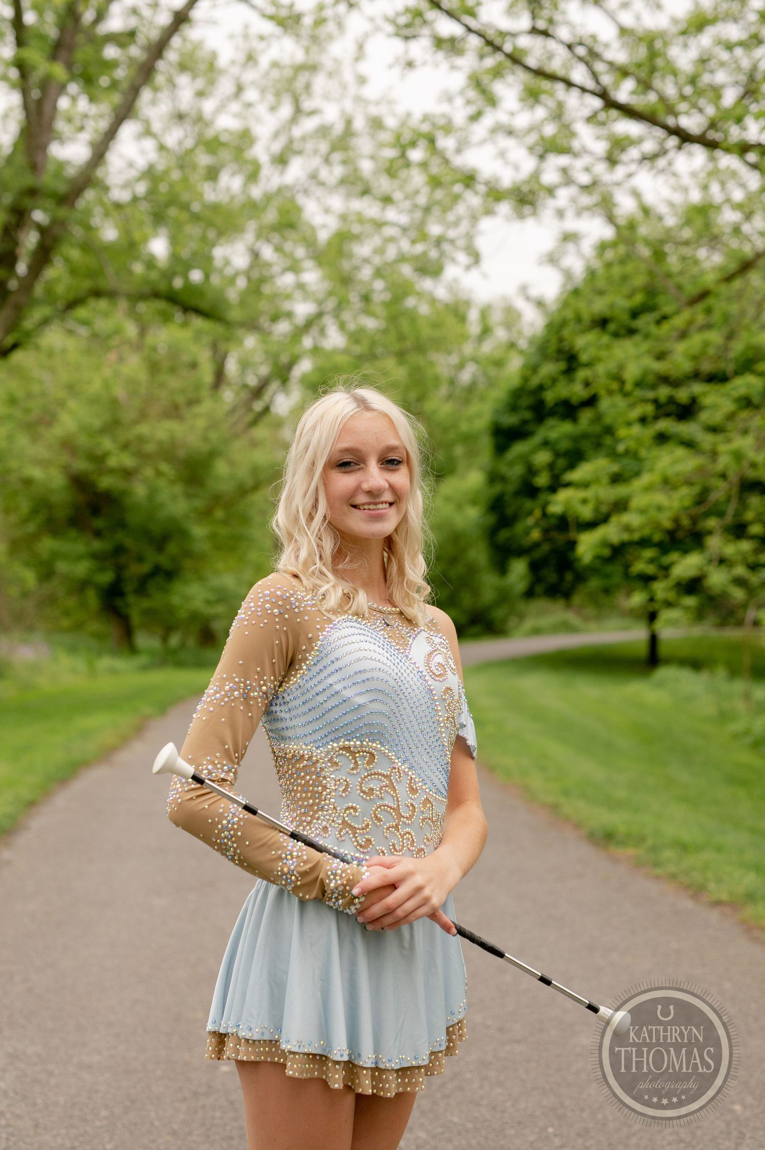 Pageant contestant in gold and blue dress holds baton while posing on park path.