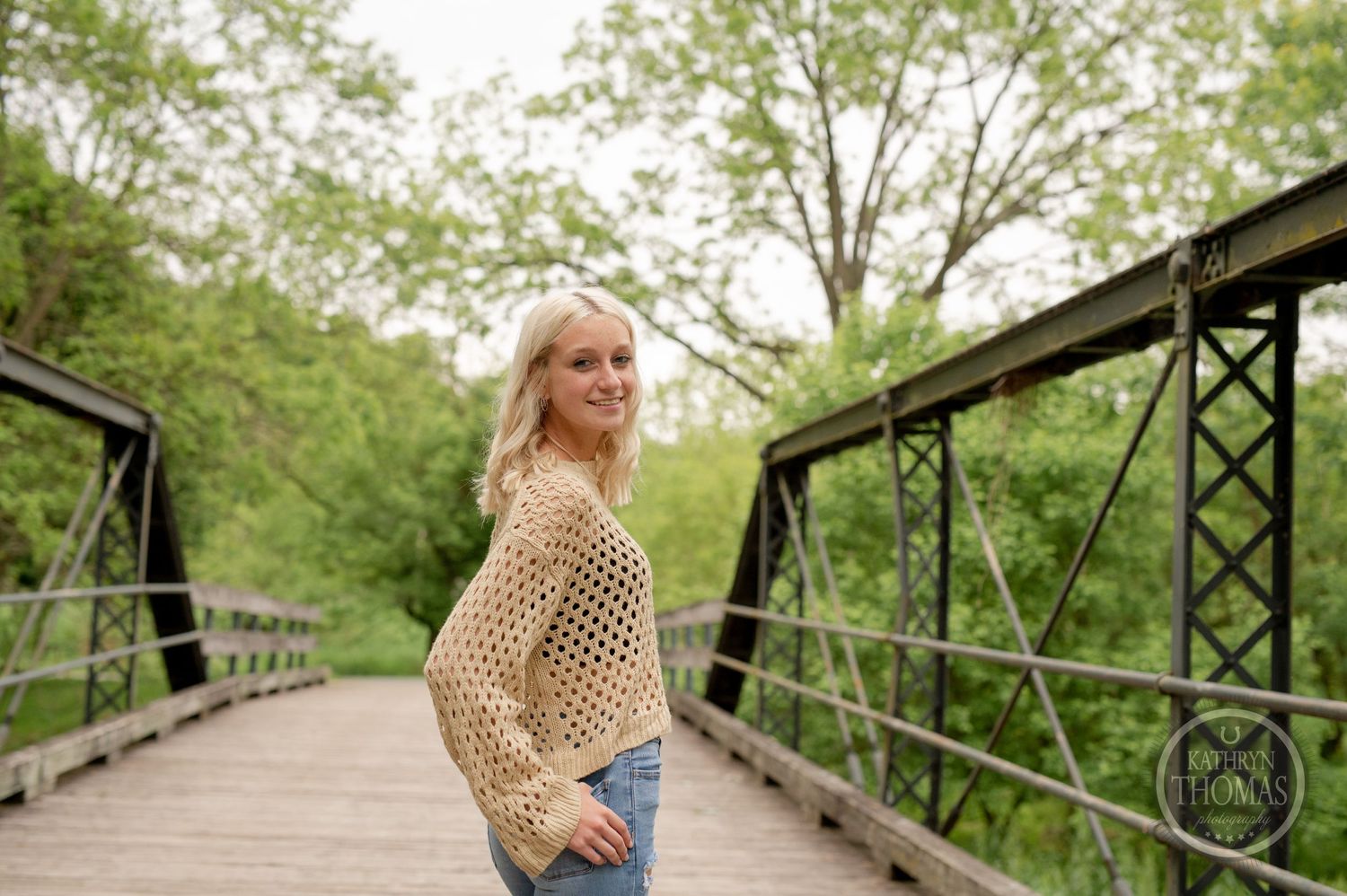 Side view portrait showcasing cream knit sweater and denim on rustic bridge with greenery.