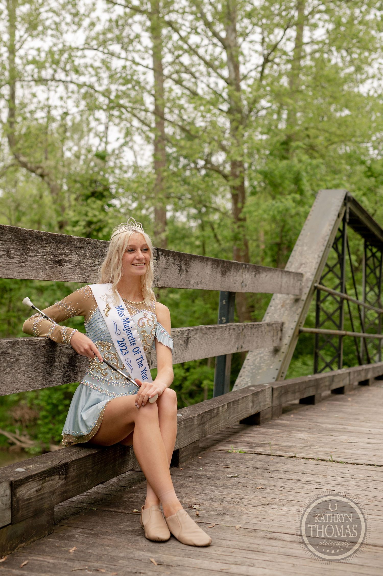 Pageant contestant sits elegantly on a wooden bridge railing while holding a scepter.