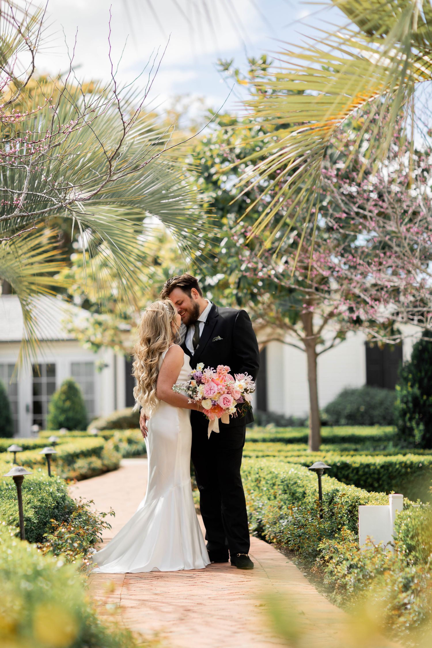 A romantic wedding couple shares tender moments in a lush garden setting with palm trees and blooming flowers in Santa Barbara.