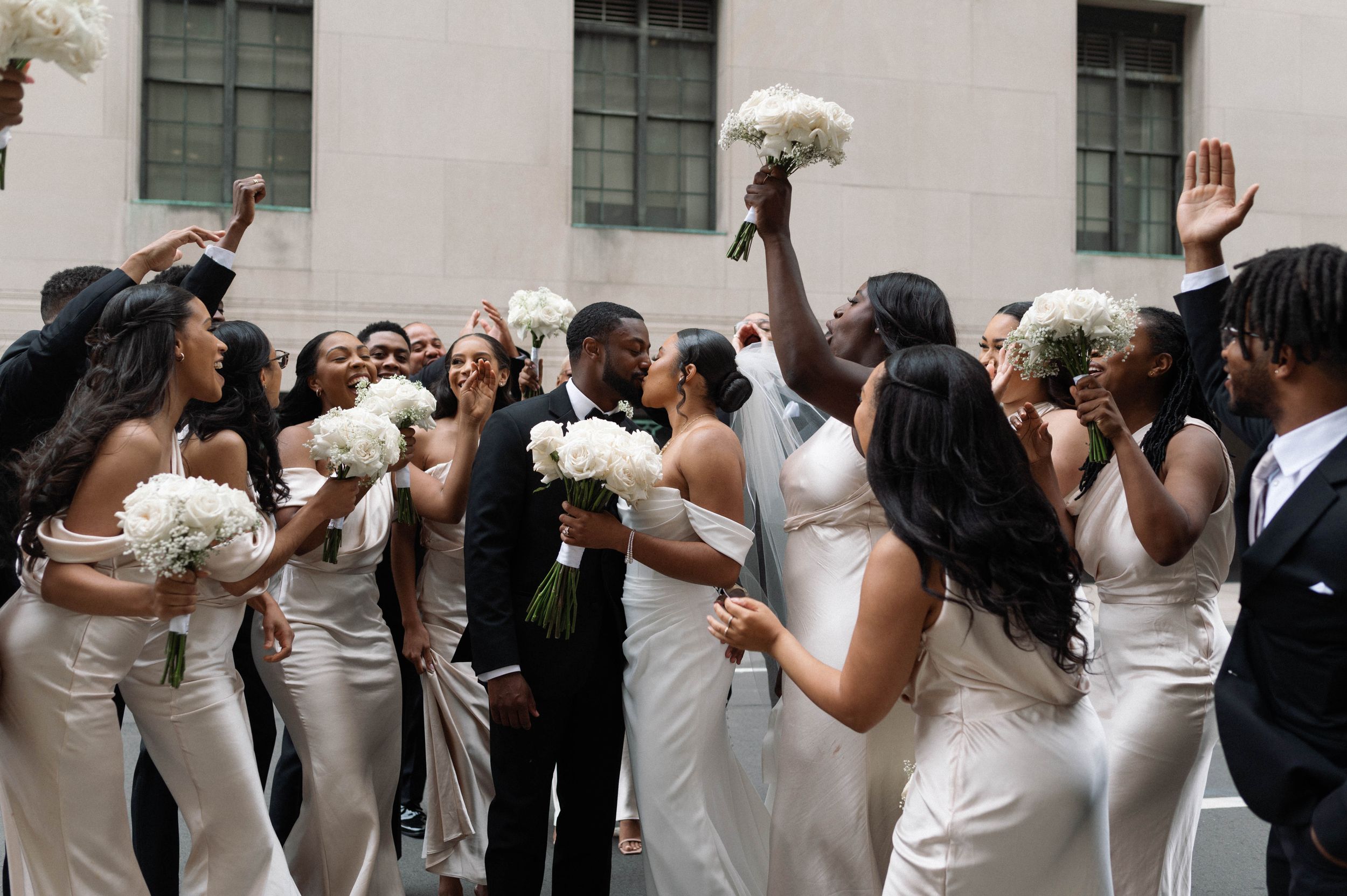 Wedding party in white dresses and black suits celebrate with raised bouquets on urban street.