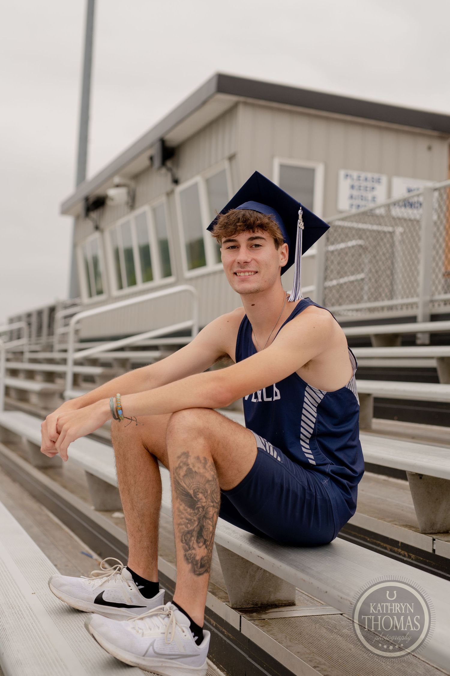 A graduate poses on stadium bleachers wearing a navy uniform and graduation cap.