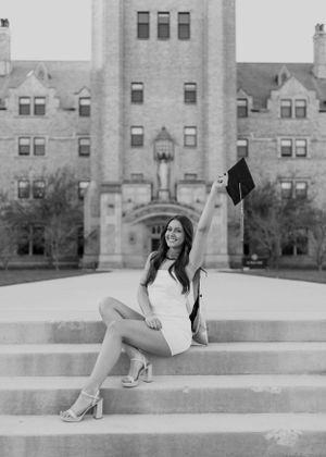 Graduate in white dress poses on grand university building steps with diploma.