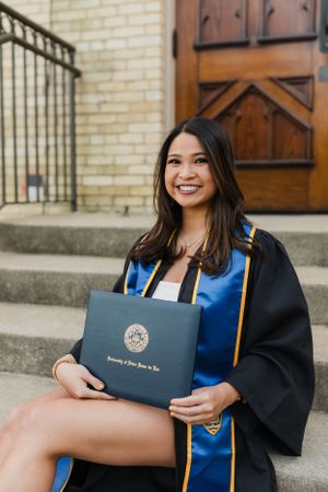Graduate holding diploma cover poses on stone steps in graduation attire.
