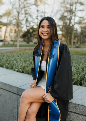 Graduate in black gown and blue stole seated outdoors during commencement.