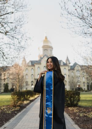 Graduate in academic regalia poses on campus path with Notre Dame's Golden Dome in background.