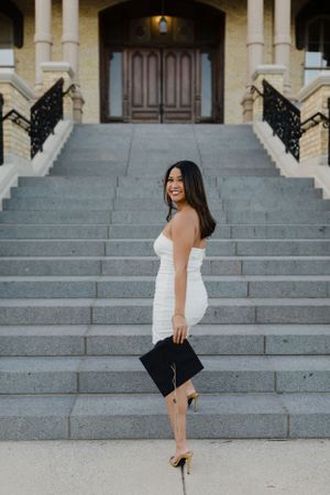 Graduate in white dress poses on stone steps during commencement photos.