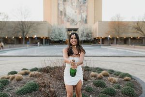 Graduate holding champagne bottle in celebratory pose by campus fountain.