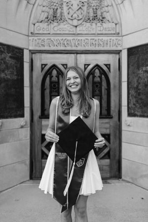 Graduate holding graduation cap in front of ornate gothic doorway at university.