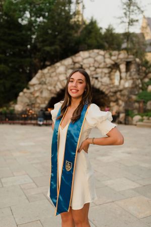 Graduate in blue stole and white dress posing by stone grotto on campus.