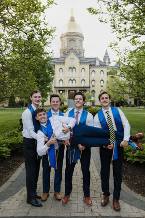 Notre Dame graduates in blue stoles posing playfully on campus pathway.