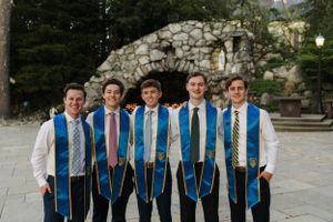 Graduates wearing blue stoles gather for photo in front of stone grotto.