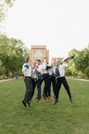 Group of graduates jumping together in celebration on campus lawn with building in background.