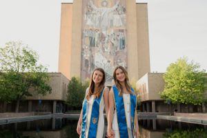 Two graduates in white dresses and blue stoles pose in front of a university building.